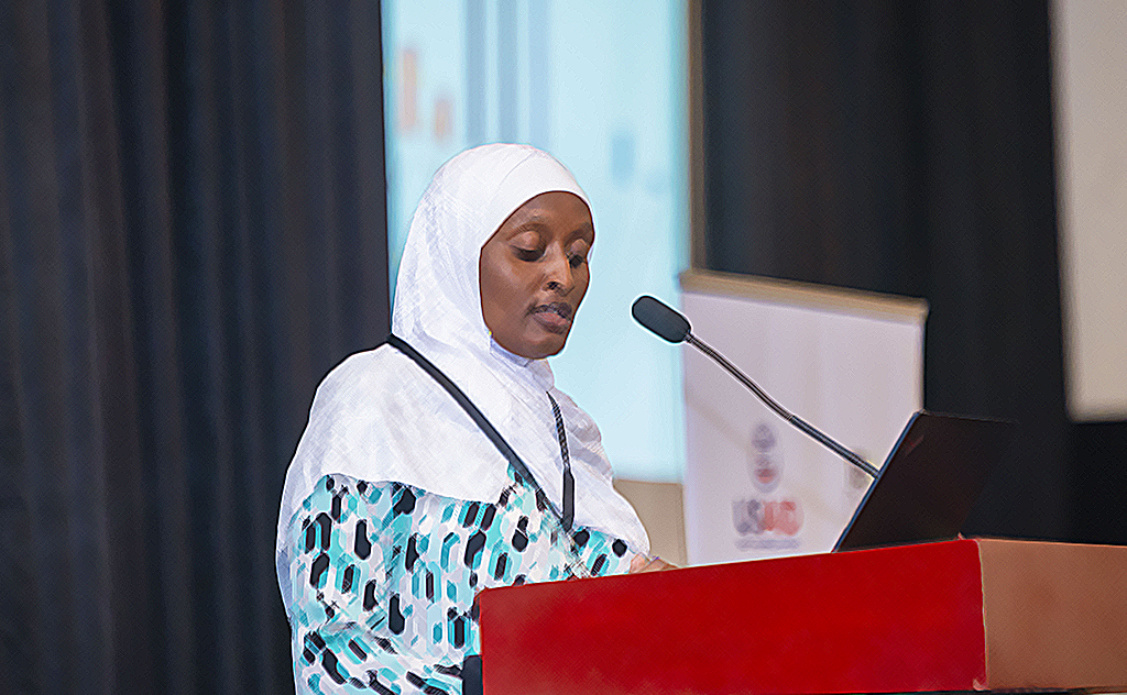 Photo of a woman in a white hijab and patterned dress speaks at a podium with a microphone, presenting at an TB regional conference hosted by USAID.